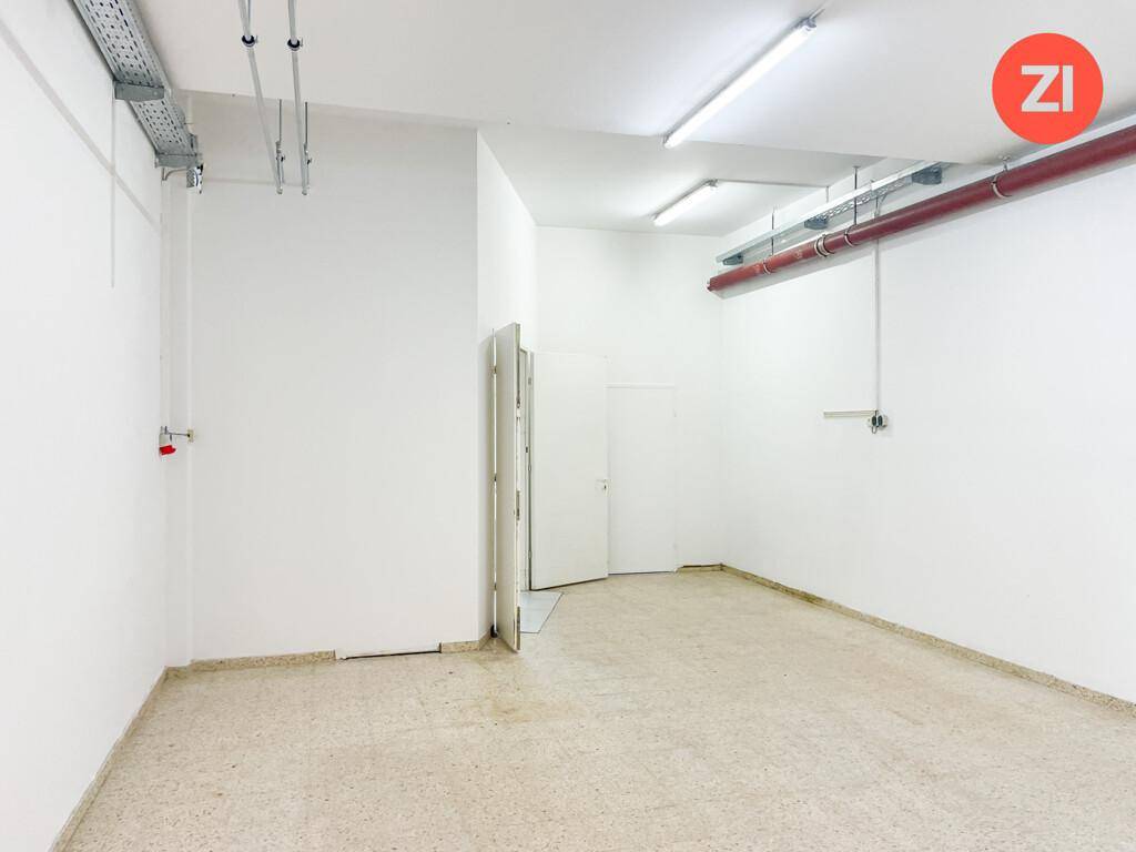 Leerer Lagerraum mit weißen Wänden, Betonboden und sichtbaren Installationen an der Decke.