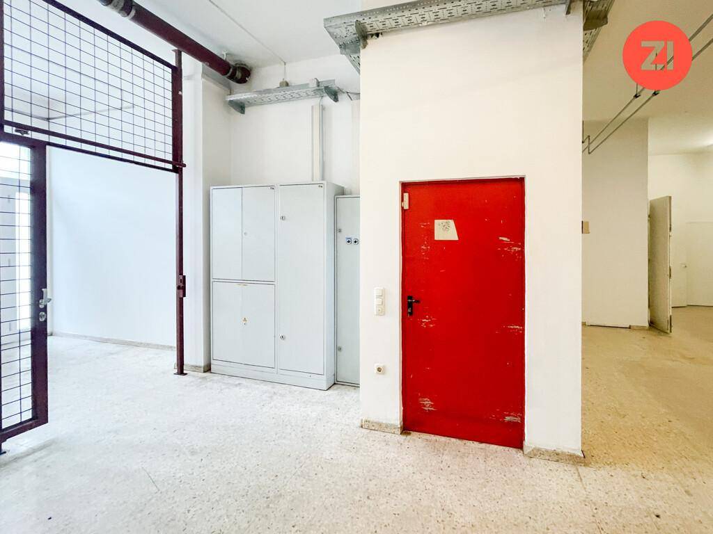 Lagerraum mit roter Tür, Metallschränken und sichtbaren Deckeninstallationen sowie Gitterabtrennung.