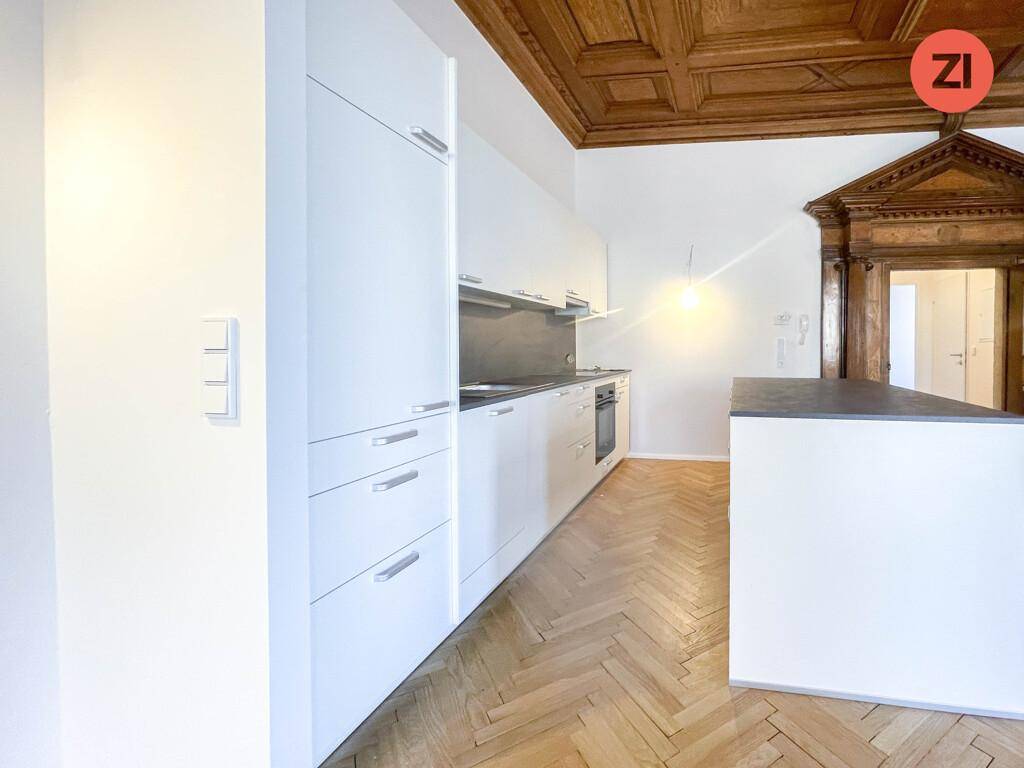 Moderne Einbauküche mit weißen Fronten und dunkler Arbeitsplatte unter einer kunstvollen Holzkassettendecke.
