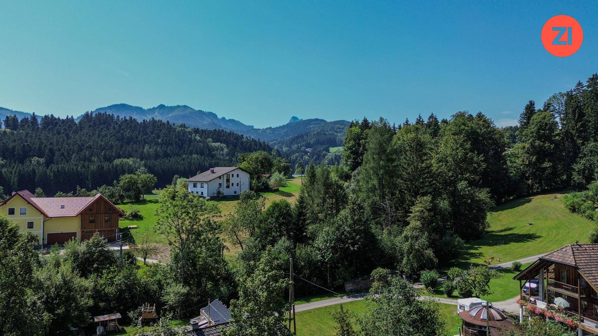 Luftaufnahme einer ländlichen Gegend mit Häusern, Wäldern und Bergen im Hintergrund unter blauem Himmel.