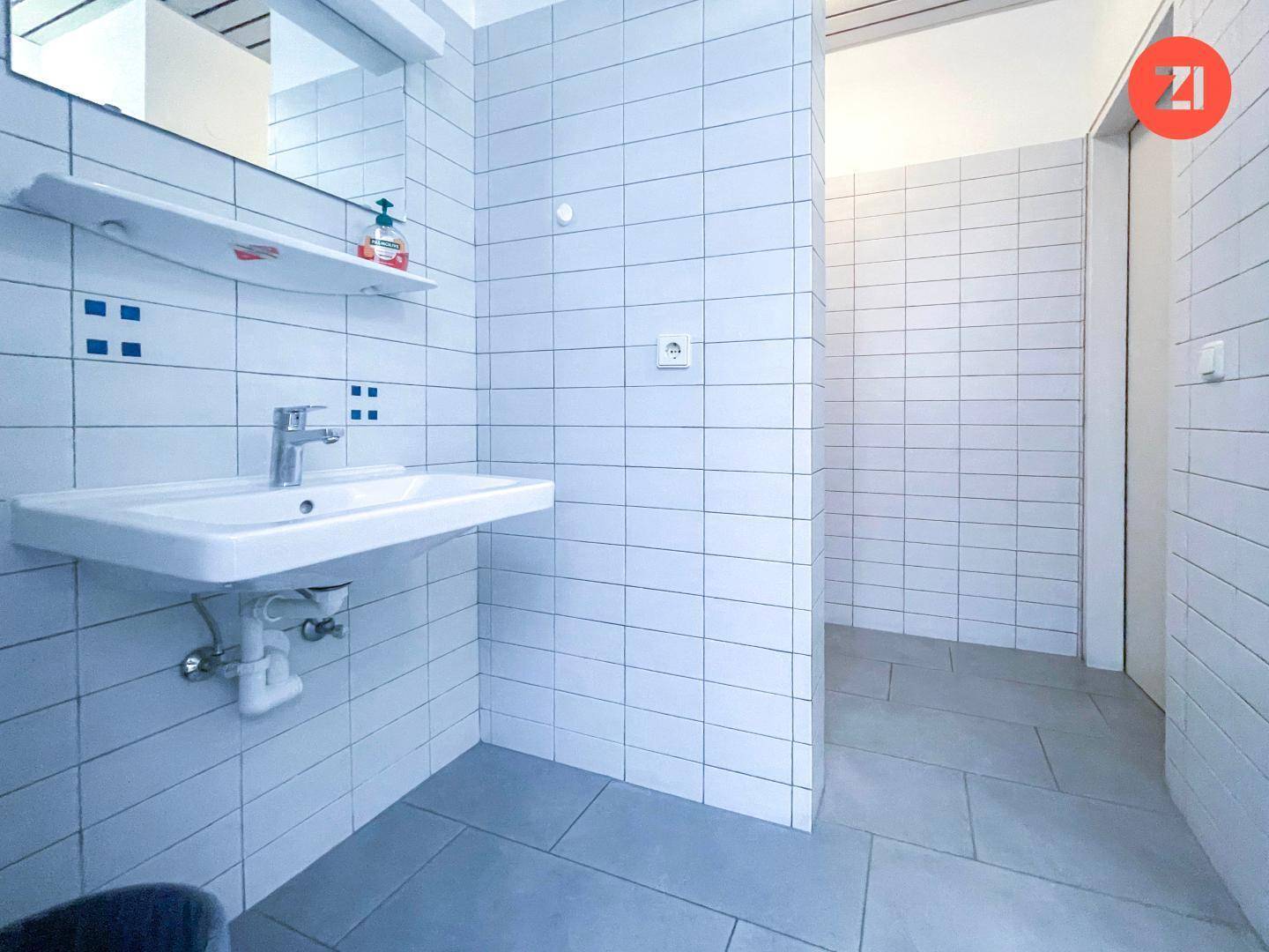 Helles Badezimmer mit weißem Waschbecken, Spiegel und weißen Fliesen an den Wänden.