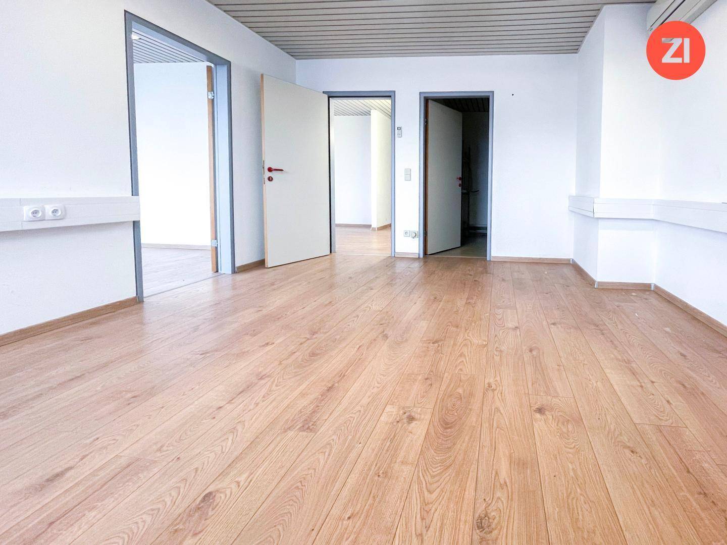 Leeres Büro mit mehreren Türen, die zu angrenzenden Räumen führen, und hellem Laminatboden.