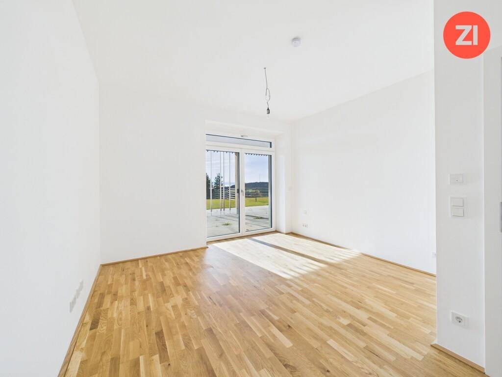 Sonniger, leerer Raum mit Holzboden und großer Glasfront, die viel Tageslicht hereinlässt.