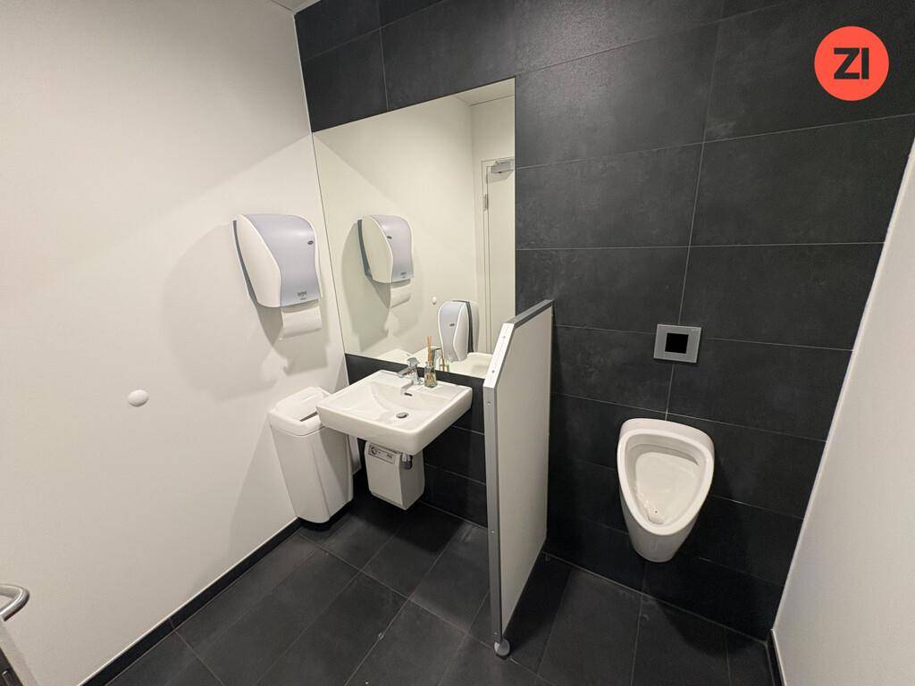 Moderne Herrentoilette mit dunklen Fliesen, Urinal, Waschbecken und großem Spiegel.