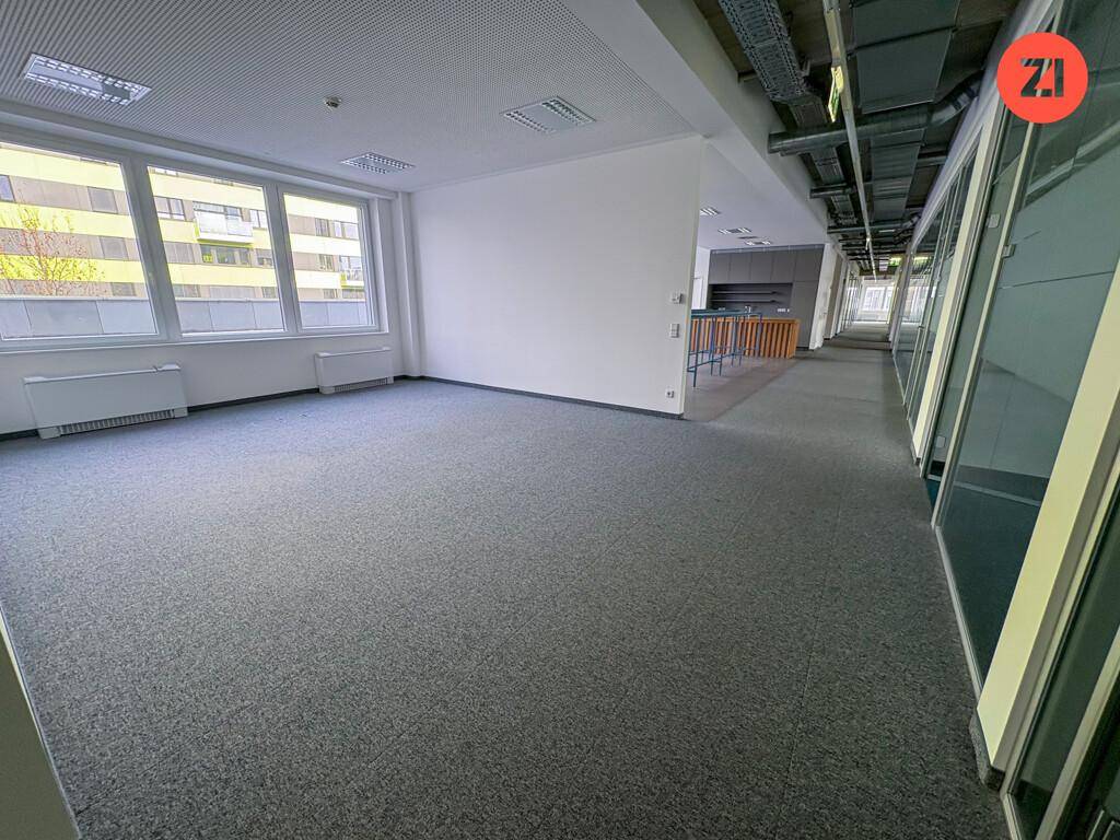Heller Büroraum mit grauem Teppichboden, großen Fenstern und Blick auf eine kleine Küchenzeile.