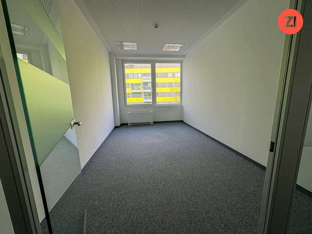 Kleiner, heller Büroraum mit grauem Teppichboden, großen Fenstern und Blick auf ein gelbes Gebäude.