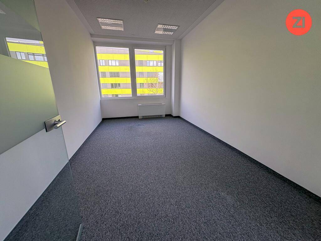 Kleiner, heller Büroraum mit grauem Teppichboden, großen Fenstern und Blick auf ein gelbes Gebäude.