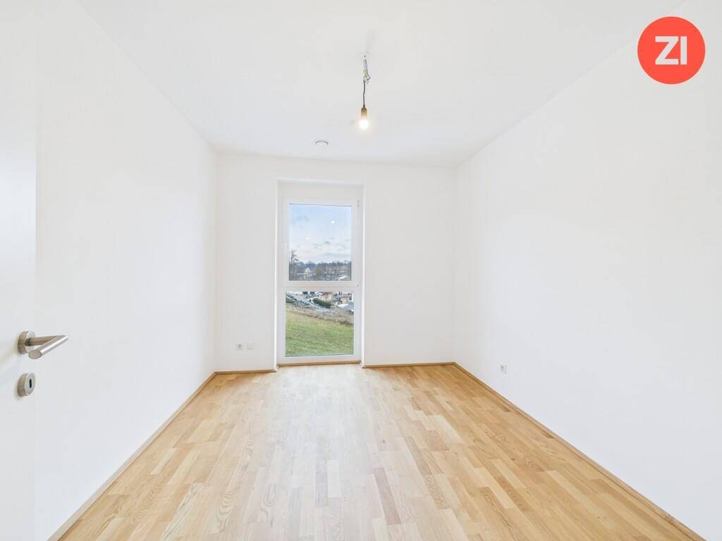 Leeres Schlafzimmer mit hellem Parkettboden und einem bodentiefen Fenster für viel Tageslicht.