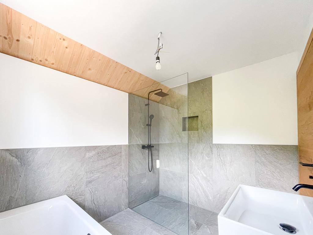 Modernes Badezimmer mit großer Dusche, Badewanne und grauen Fliesen, unter einer Holzdecke.