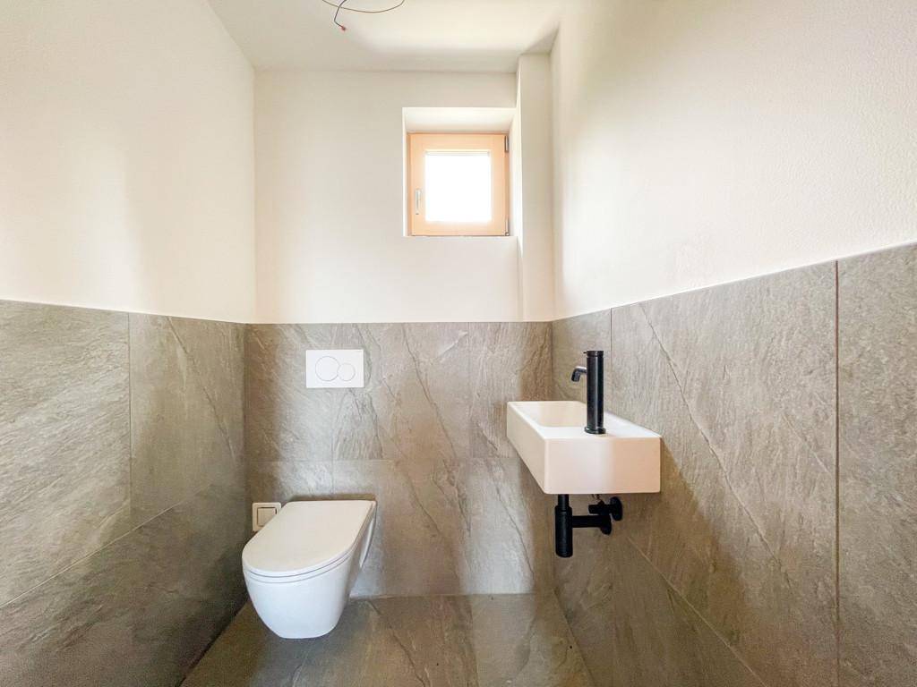 Modernes Gäste-WC mit grauen Fliesen, wandhängendem WC und kleinem Waschbecken mit schwarzer Armatur.