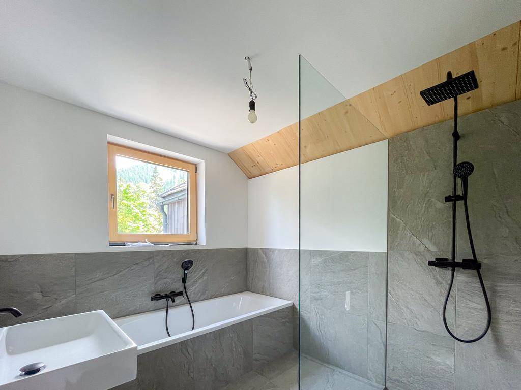 Modernes Badezimmer mit Badewanne, Dusche und grauen Fliesen, unter einer Holzdecke.