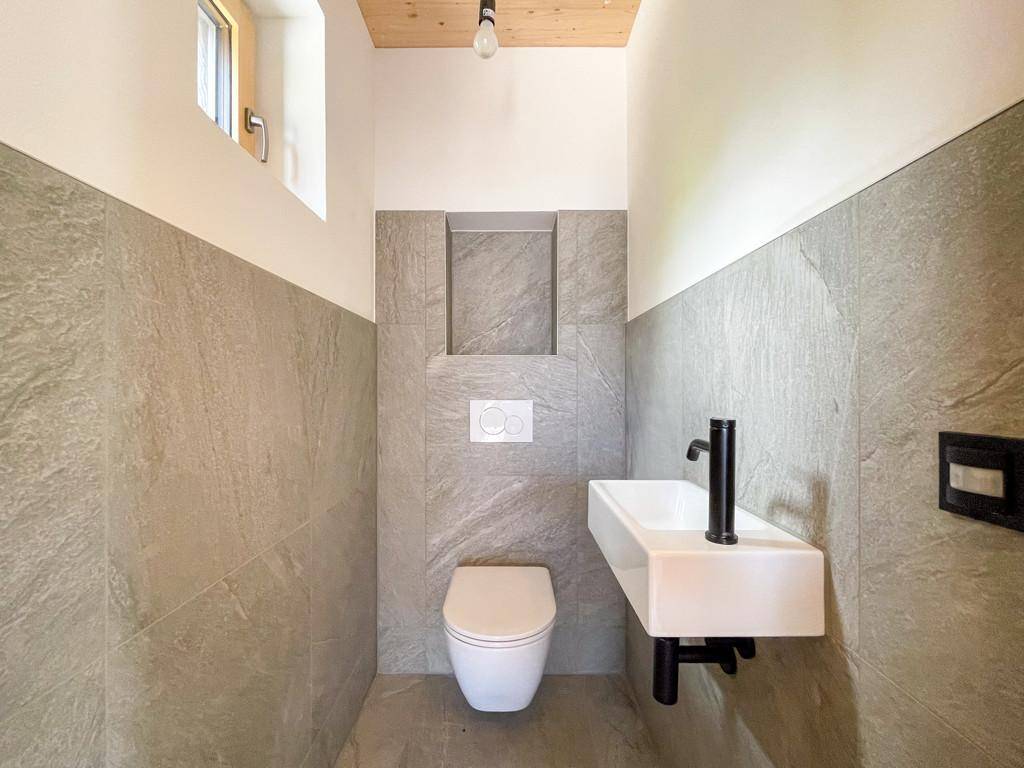 Modernes Gäste-WC mit grauen Fliesen, wandhängendem WC und kleinem Waschbecken mit schwarzer Armatur.