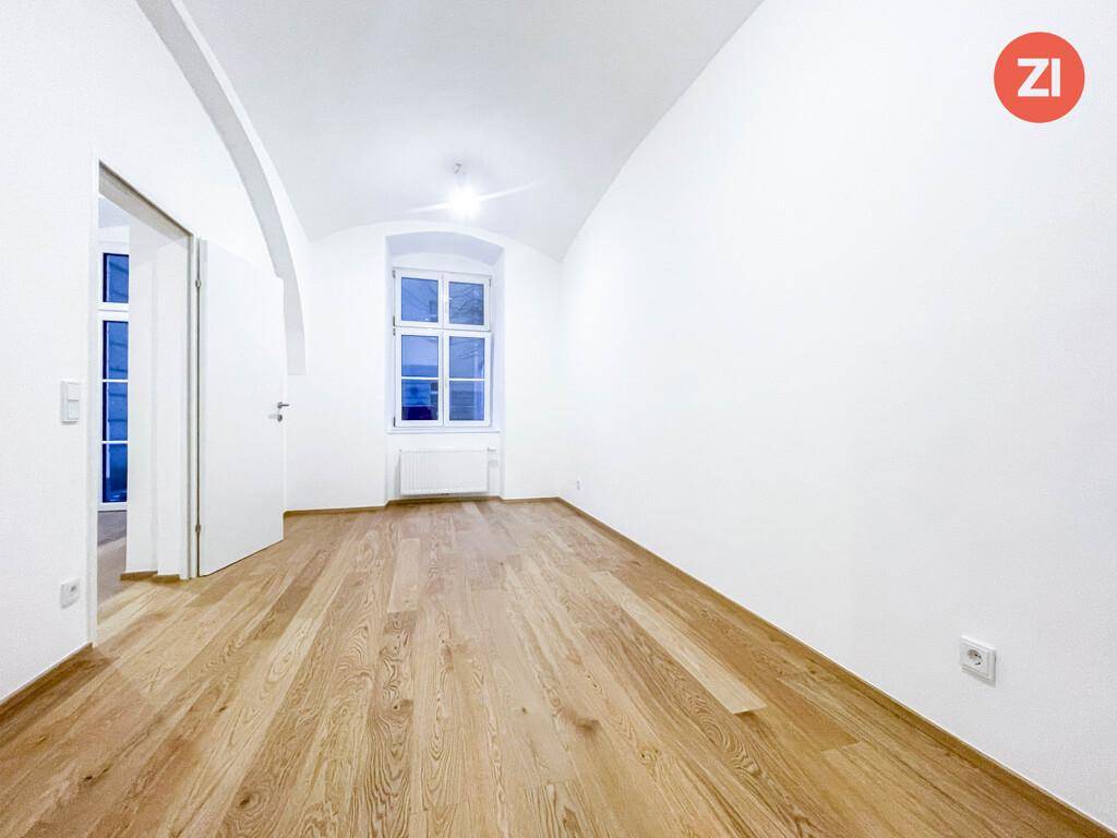 Helles Zimmer mit Holzboden, einem großen Fenster und gewölbter Decke.