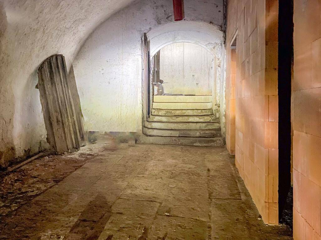 Gewölbter Kellergang mit Stufen, die zu einem Torbogen führen, und einer Mischung aus alten und neuen Baumaterialien.