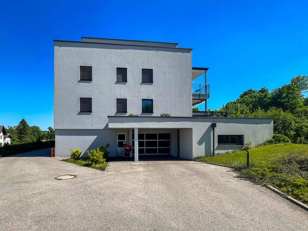 Modernes graues Gebäude mit integrierter Garage, Balkon und gepflegter Auffahrt unter blauem Himmel.