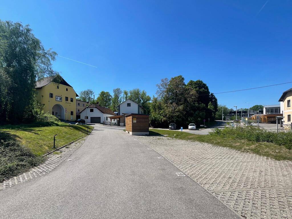 Großer Außenparkplatz mit Schotterflächen und einer Zufahrt, umgeben von Bäumen und Gebäuden.