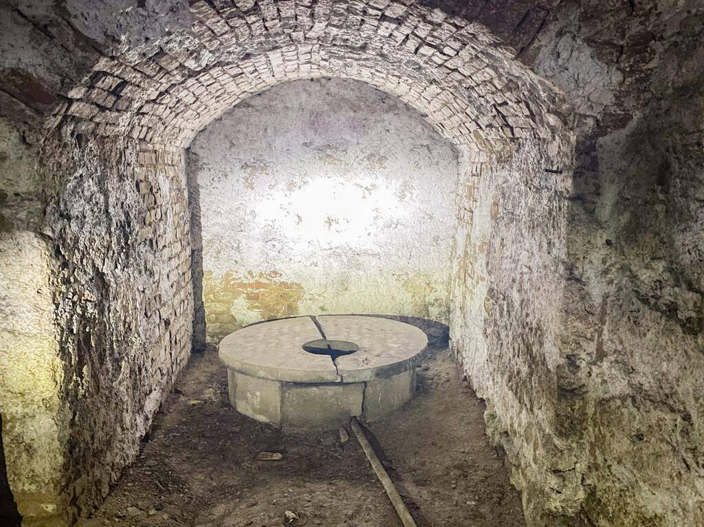 Kleiner, gewölbter Kellerraum mit Ziegelwänden und einem runden Steinobjekt in der Mitte, möglicherweise ein alter Brunnen.