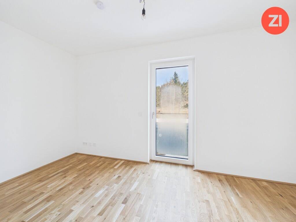 Leeres Schlafzimmer mit Holzboden und einem hohen Fenster, das viel Licht hereinlässt.