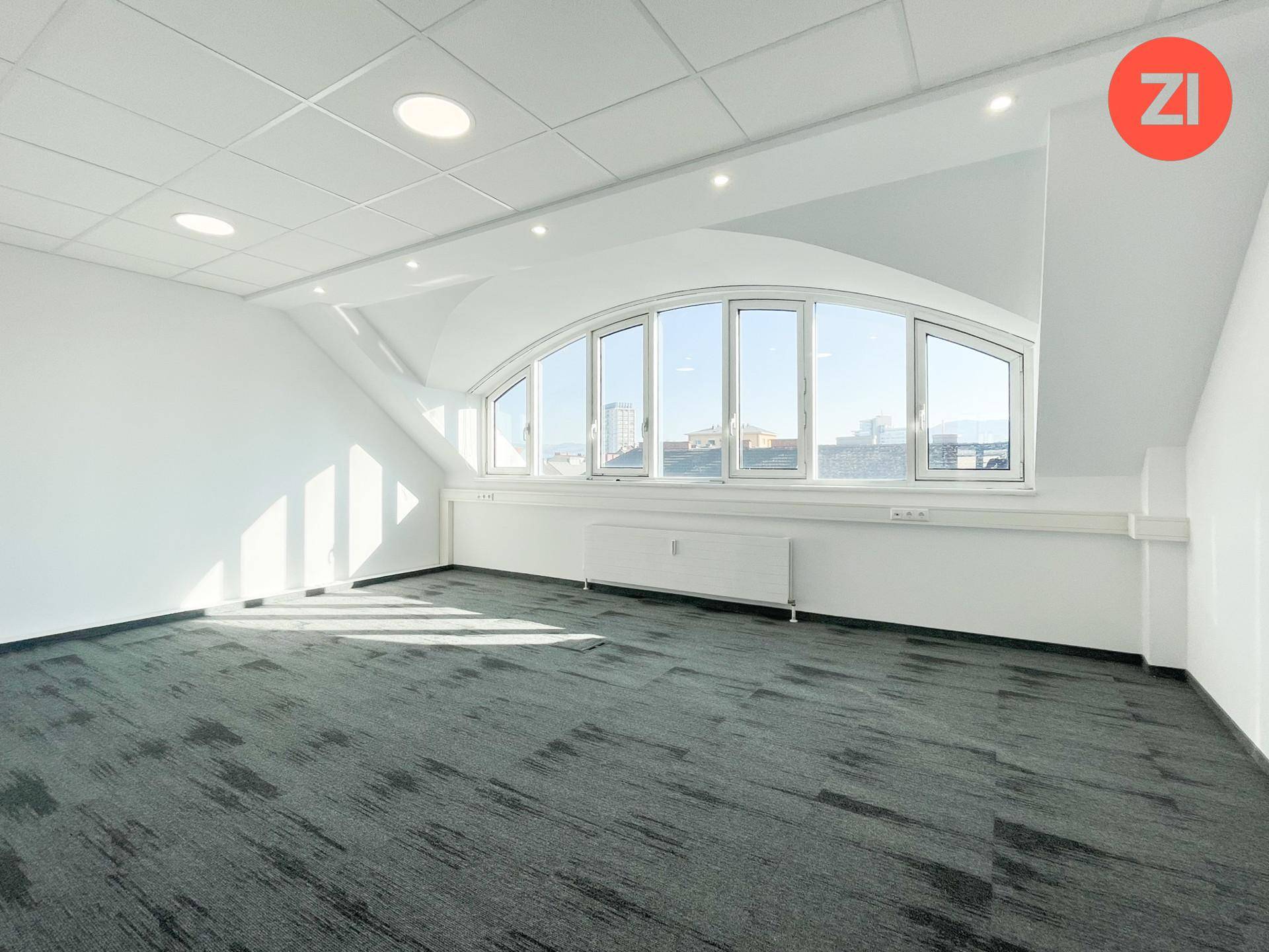 Helles Büro mit Teppichboden und einer großen, bogenförmigen Fensterfront, die viel Tageslicht hereinlässt.