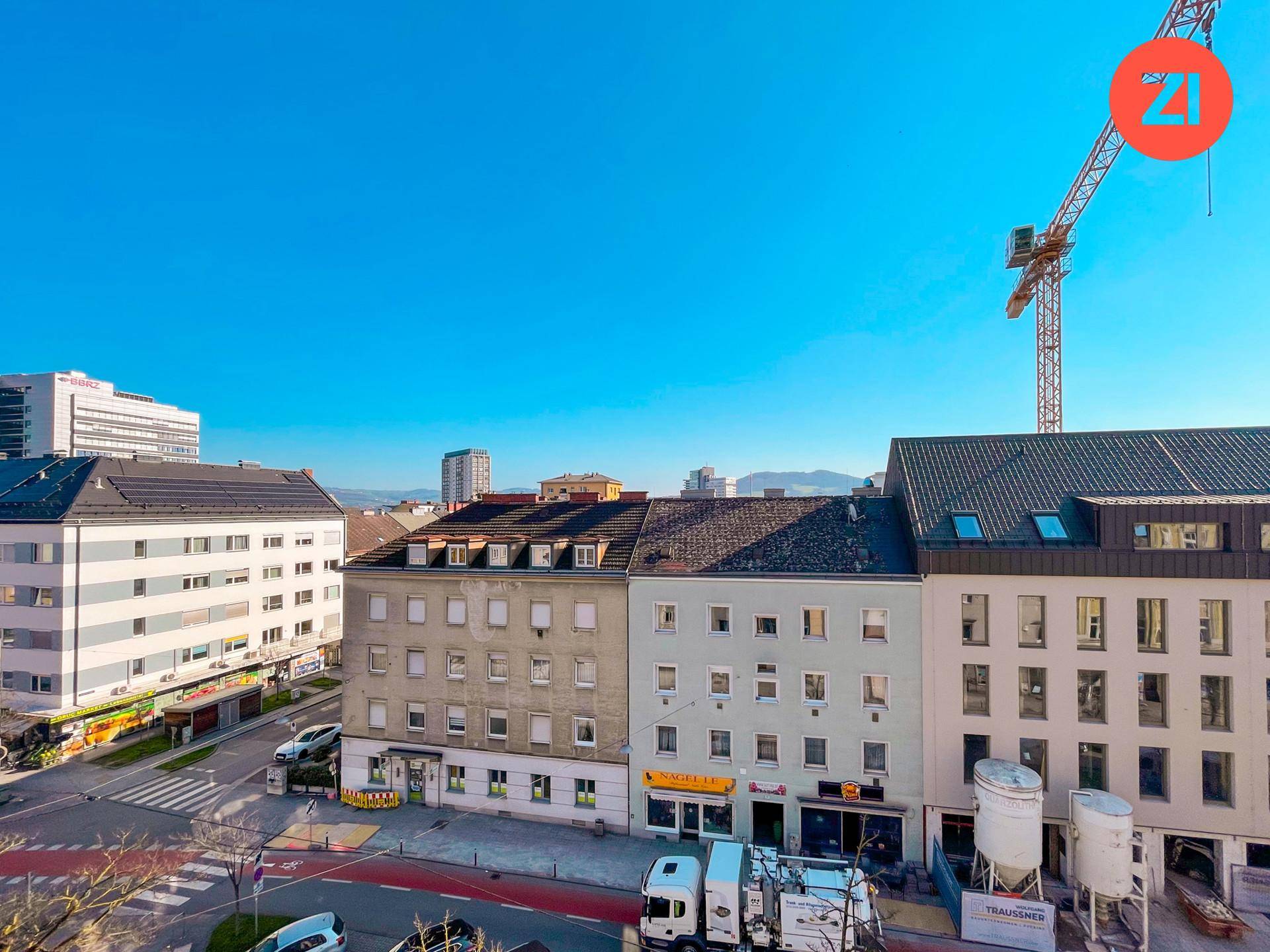 Stadtansicht mit Gebäuden, einer Baustelle und Bergen im Hintergrund unter klarem Himmel.