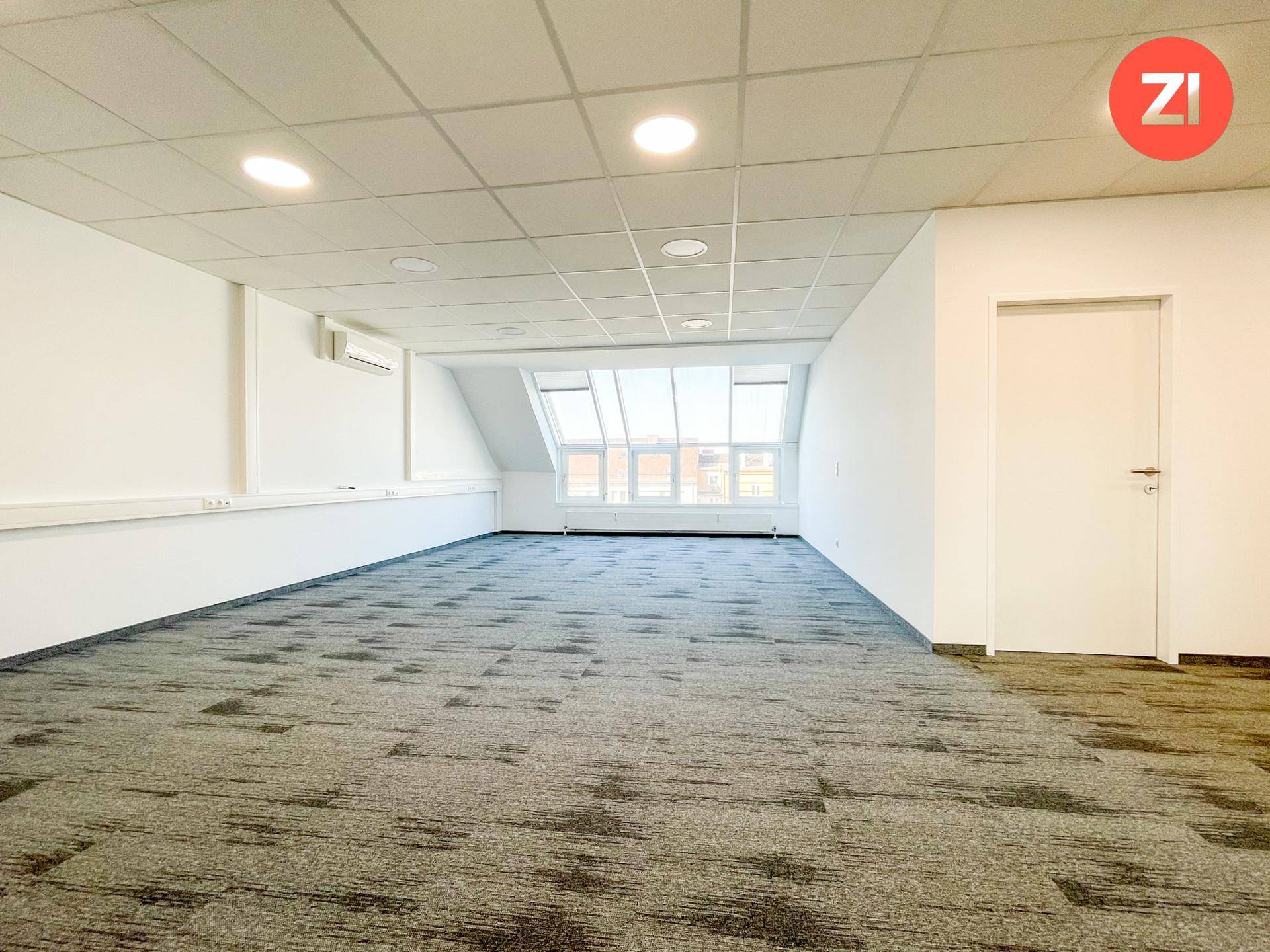 Langer Büroraum mit Teppichboden, Klimaanlage und einer Fensterfront im Dachgeschoss.