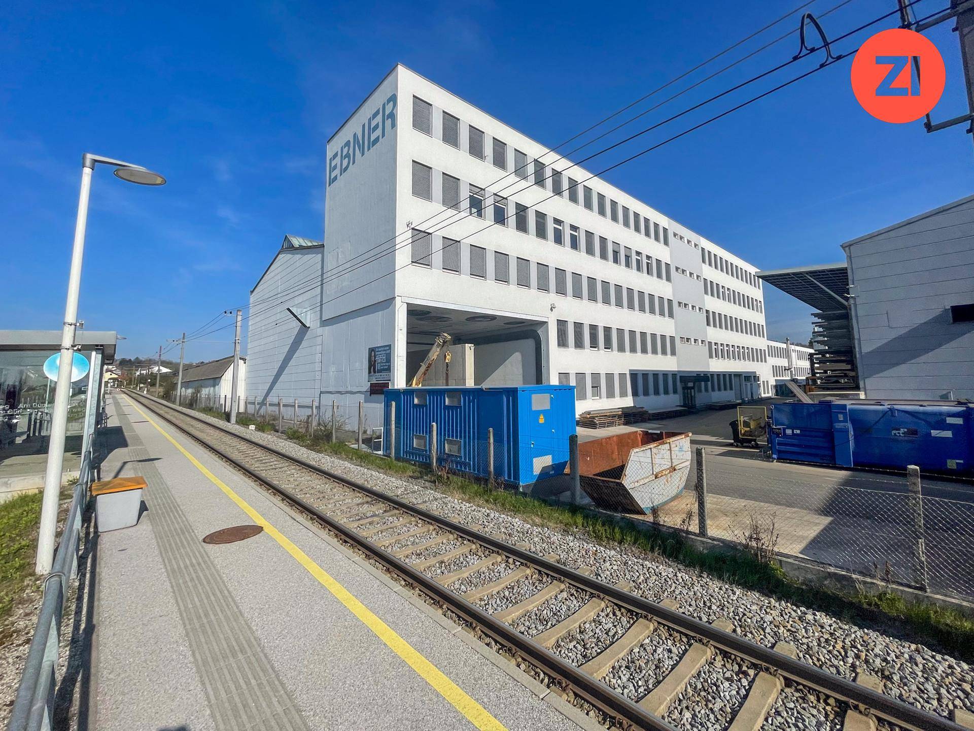 Außenansicht eines großen weißen Industriegebäudes neben Bahngleisen mit blauen Containern.
