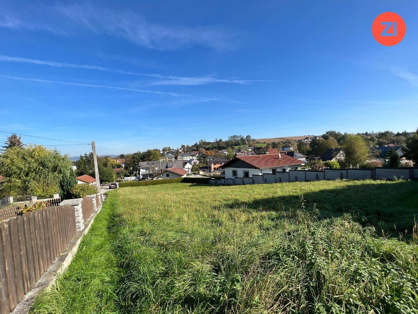 Offenes Grundstück mit grüner Wiese, umgeben von einem Holzzaun und Wohnhäusern in der Ferne.