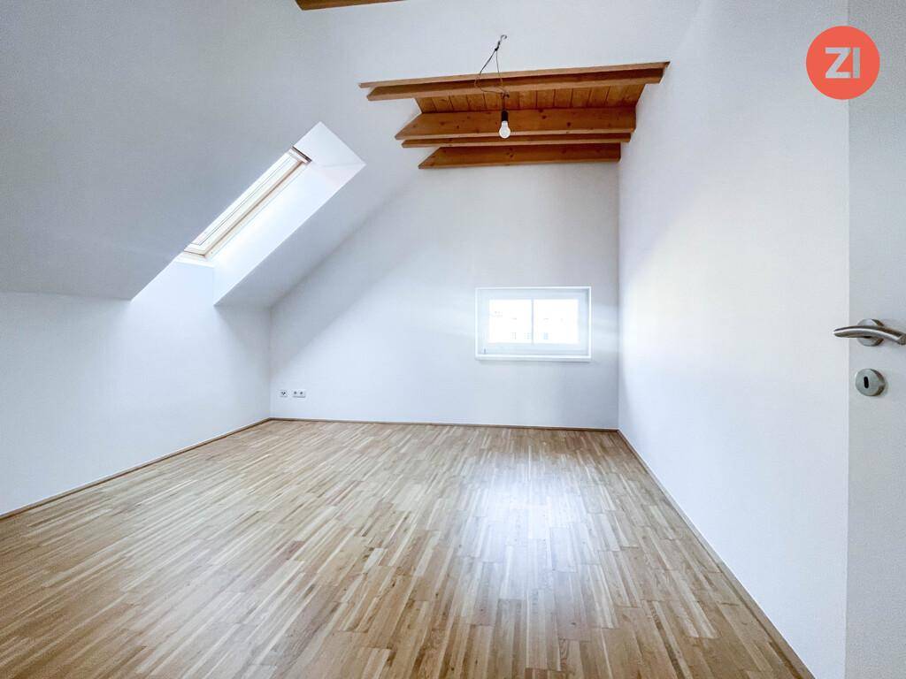 Helles Zimmer im Dachgeschoss mit Holzboden, Dachfenster und einem weiteren Fenster, das viel Licht hereinlässt.
