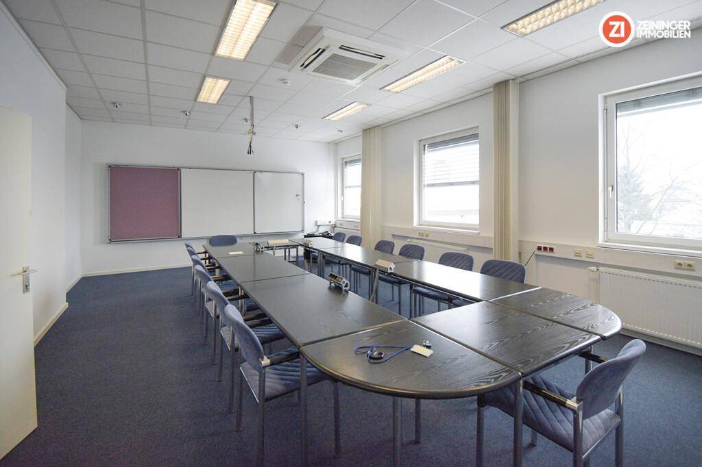 Konferenzraum mit großem U-förmigem Tisch, vielen Stühlen, Teppichboden und Fenstern mit Jalousien.