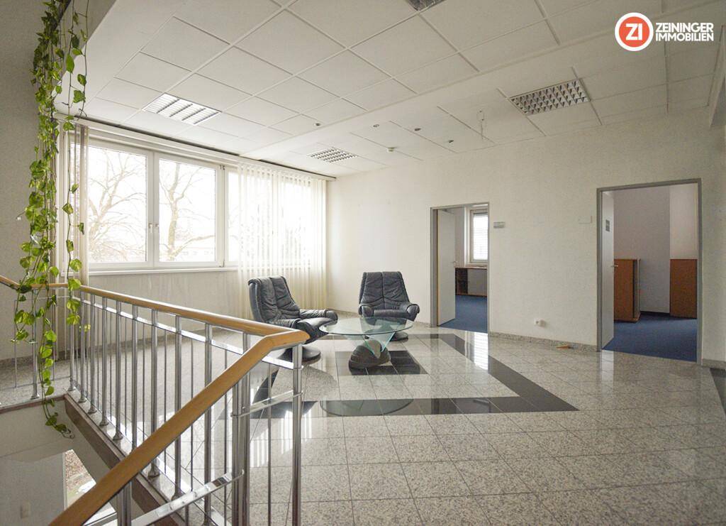 Empfangsbereich mit Fliesenboden, kleiner Sitzecke und einer Treppe mit Metallgeländer, hell durch große Fenster.