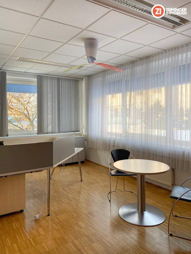 Helles Büro mit Holzfußboden, großen Fenstern mit Jalousien und einem Deckenventilator.