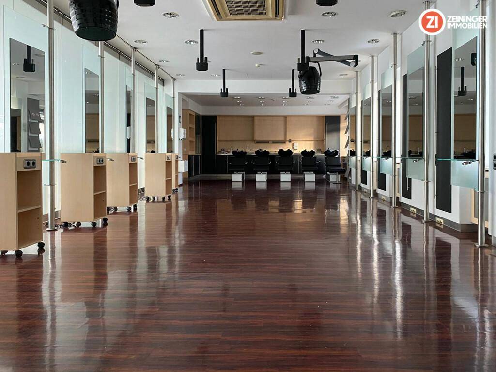 Innenansicht eines Friseursalons mit dunklem Holzfußboden, mehreren Styling-Stationen und Waschbecken im Hintergrund.