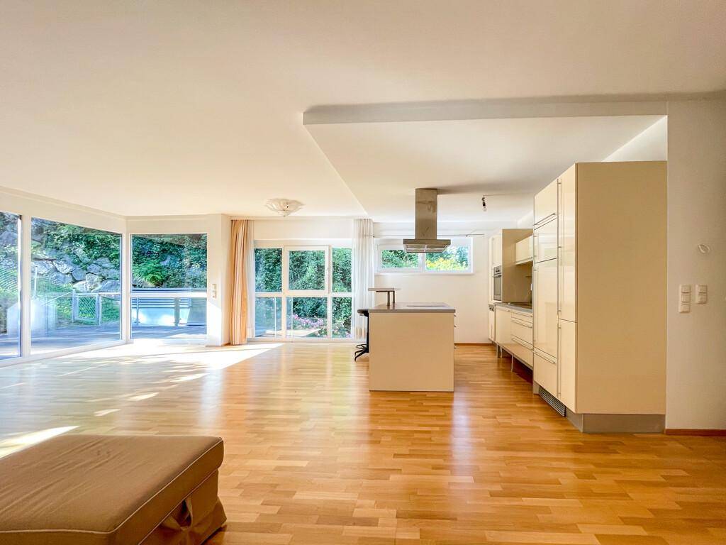 Weitläufiger Wohn- und Essbereich mit offener Küche, Parkettboden und Zugang zu den großen Fensterfronten.