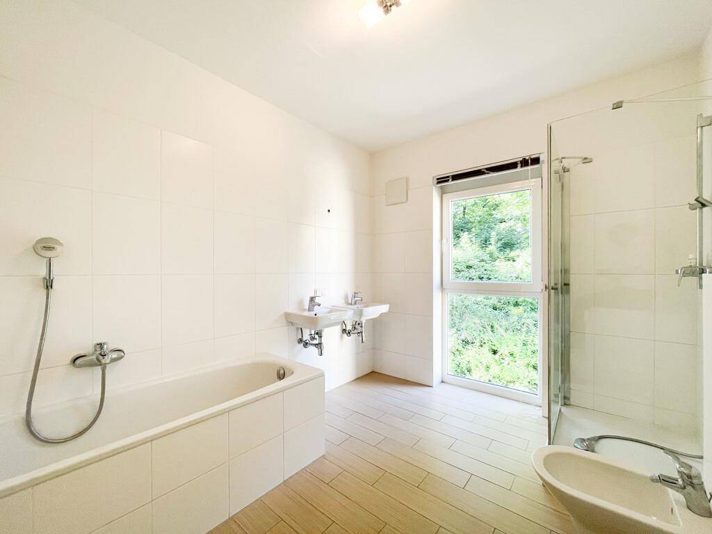 Geräumiges Badezimmer mit weiß gefliesten Wänden, Badewanne, Dusche, Doppelwaschbecken und Fenster mit Grünblick.