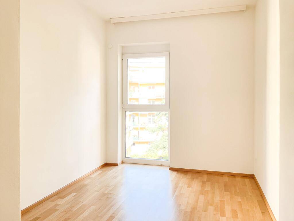 Leeres Zimmer mit hellem Parkettboden und einem Fenster, das den Blick auf die umliegenden Gebäude freigibt.