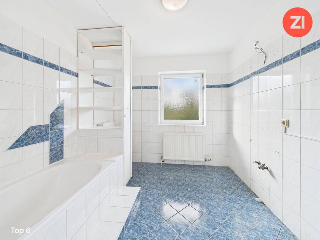 Badezimmer mit weißen und blauen Fliesen, Badewanne und einem Fenster, das Tageslicht hereinlässt.