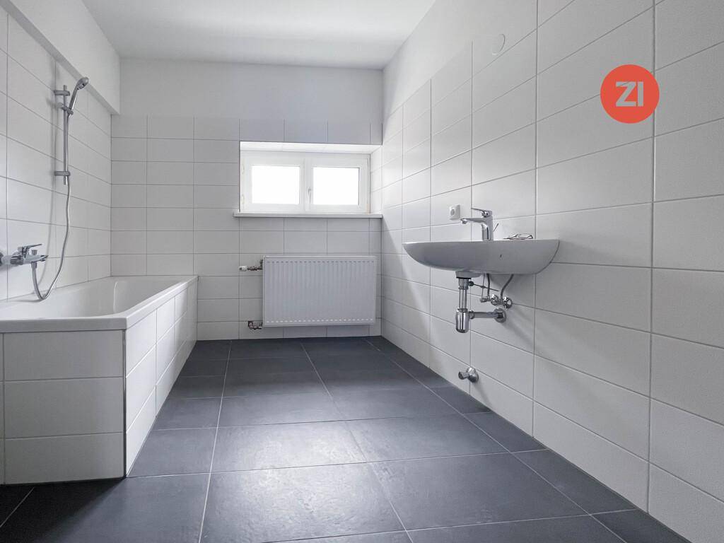 Modern gefliestes Badezimmer mit Badewanne, Waschbecken und Fenster für natürliche Belüftung.