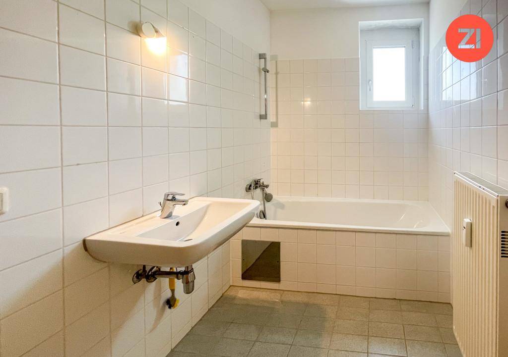 Badezimmer mit Badewanne, Waschbecken, weißen Fliesen und einem Fenster für Tageslicht.