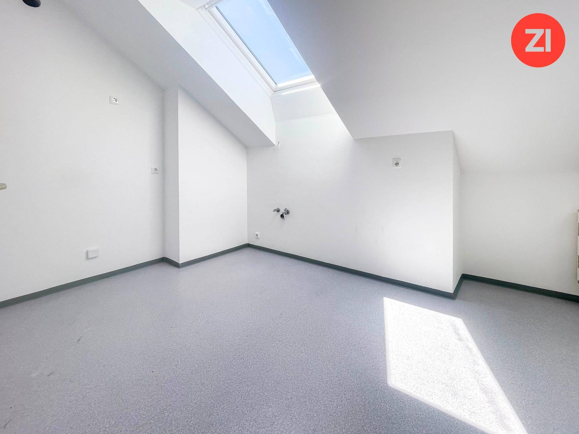 Leerer Raum mit grauem Boden, Dachschräge und einem Dachfenster, vorbereitet für Installationen.