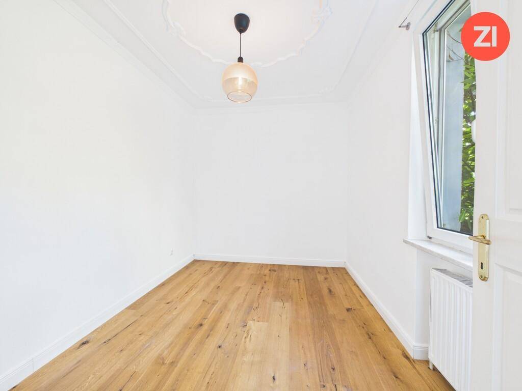 Helles Zimmer mit Holzboden, Stuckdecke und einem Fenster mit Blick ins Grüne.