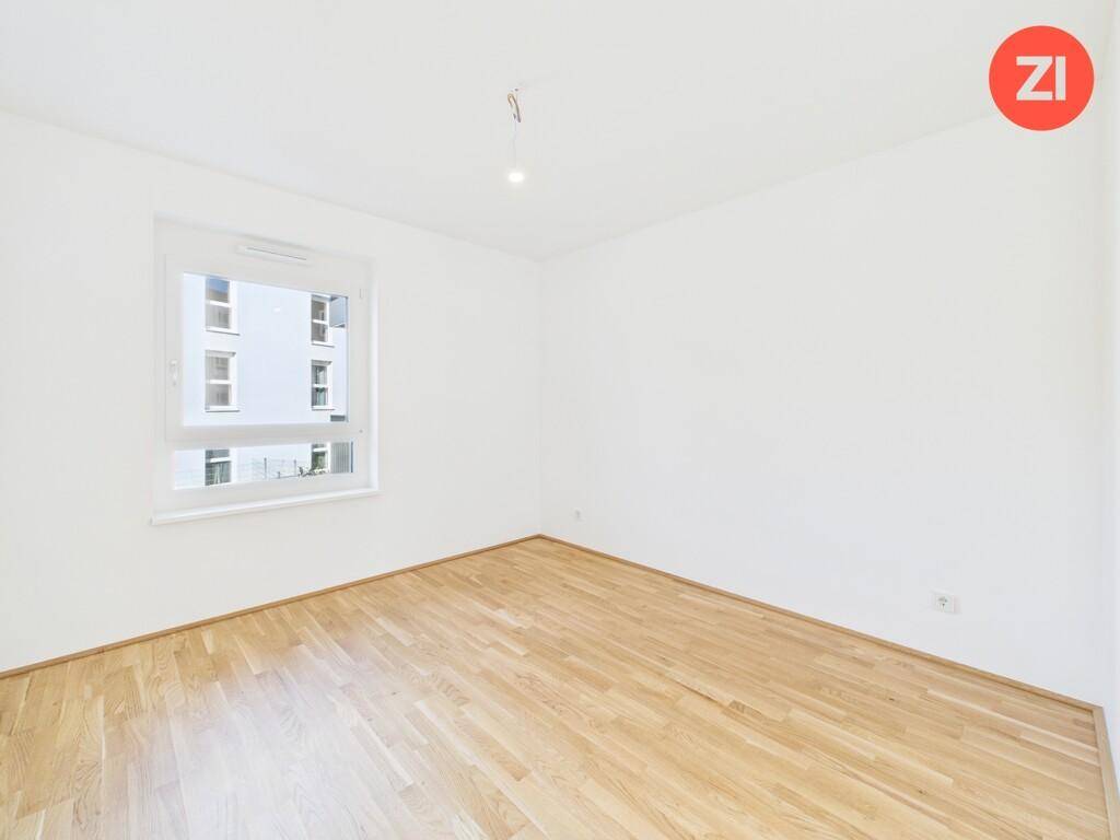 Leeres Schlafzimmer mit hellem Parkettboden, weißen Wänden und einem Fenster mit Blick auf andere Gebäude.