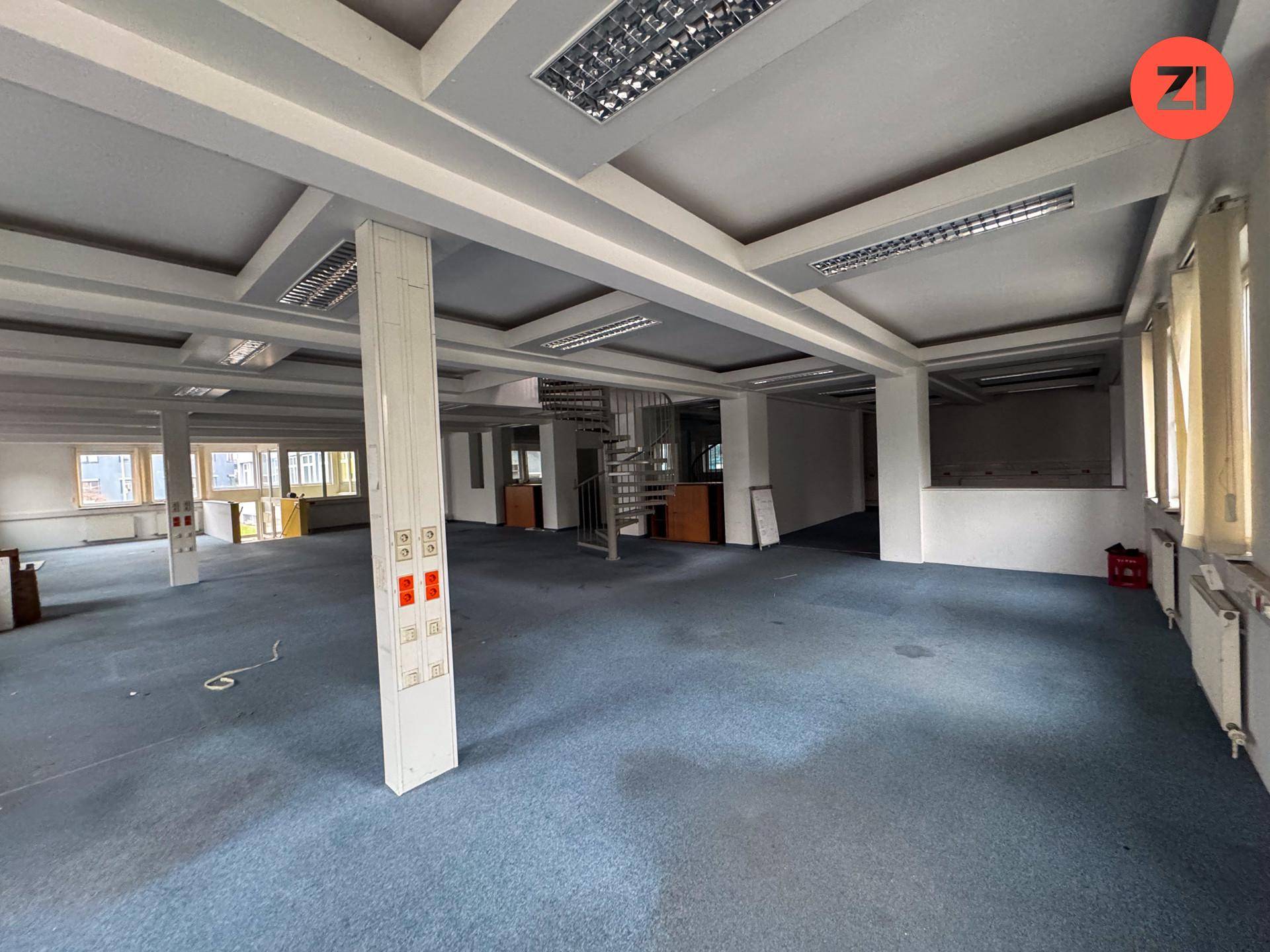 Großes, offenes Büro mit Teppichboden, Rasterdeckenbeleuchtung und einer Wendeltreppe in der Mitte.