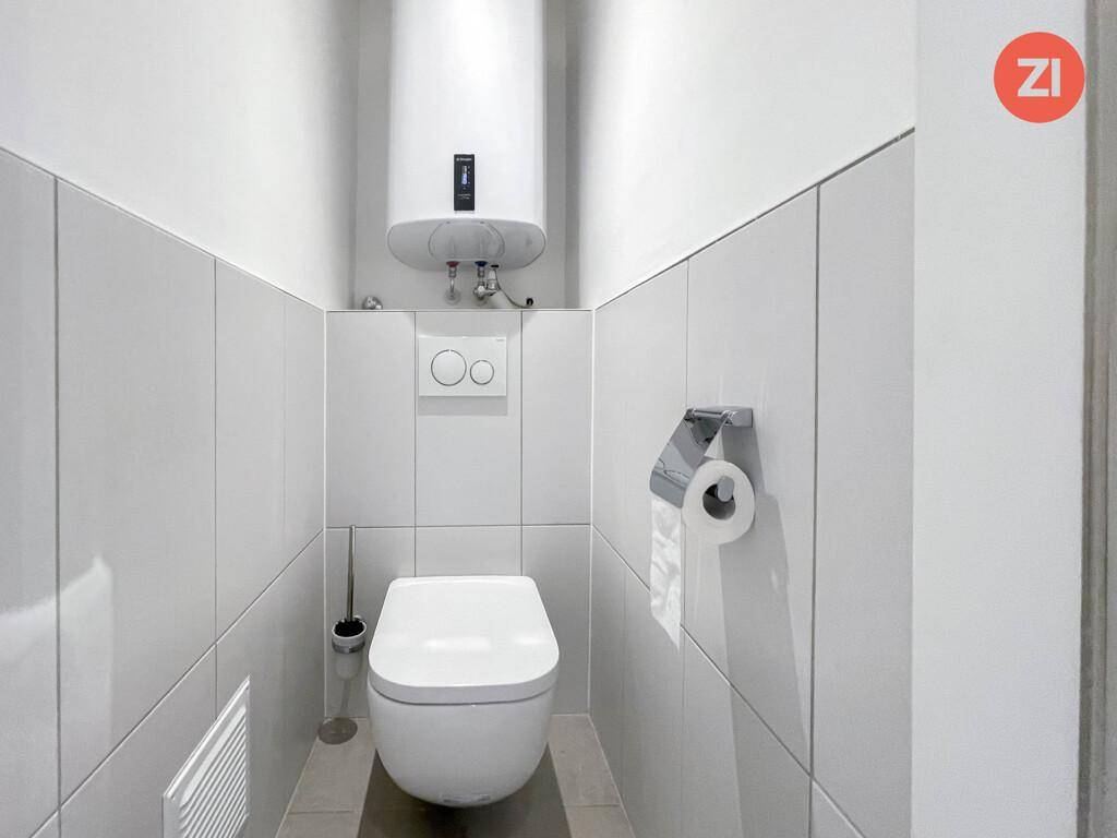 Separates WC mit wandhängender Toilette, weißen Fliesen und Warmwasserboiler an der Wand.