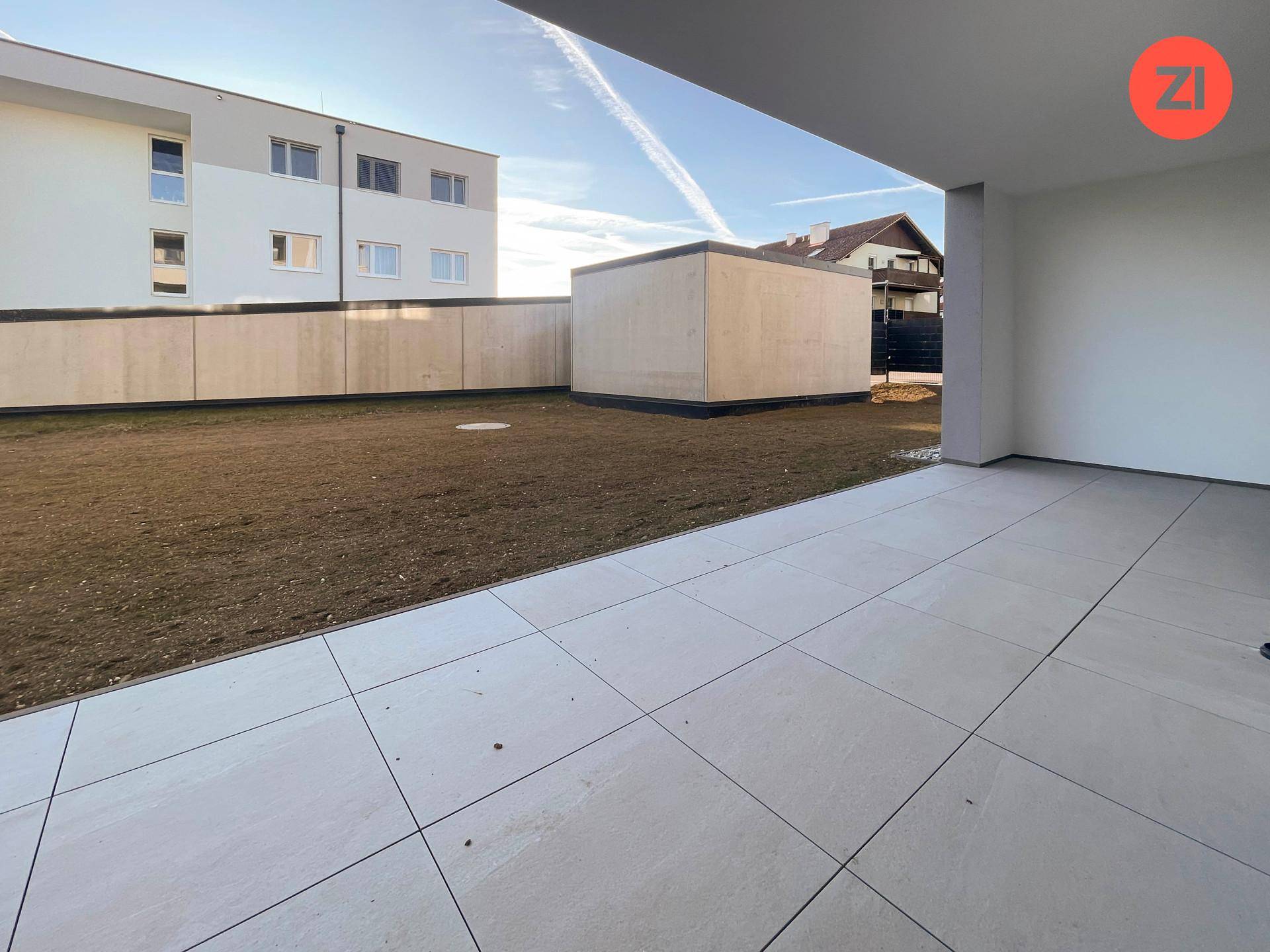 Unmöblierte Terrasse mit Fliesenboden und angrenzender Rasenfläche vor einem Neubau.