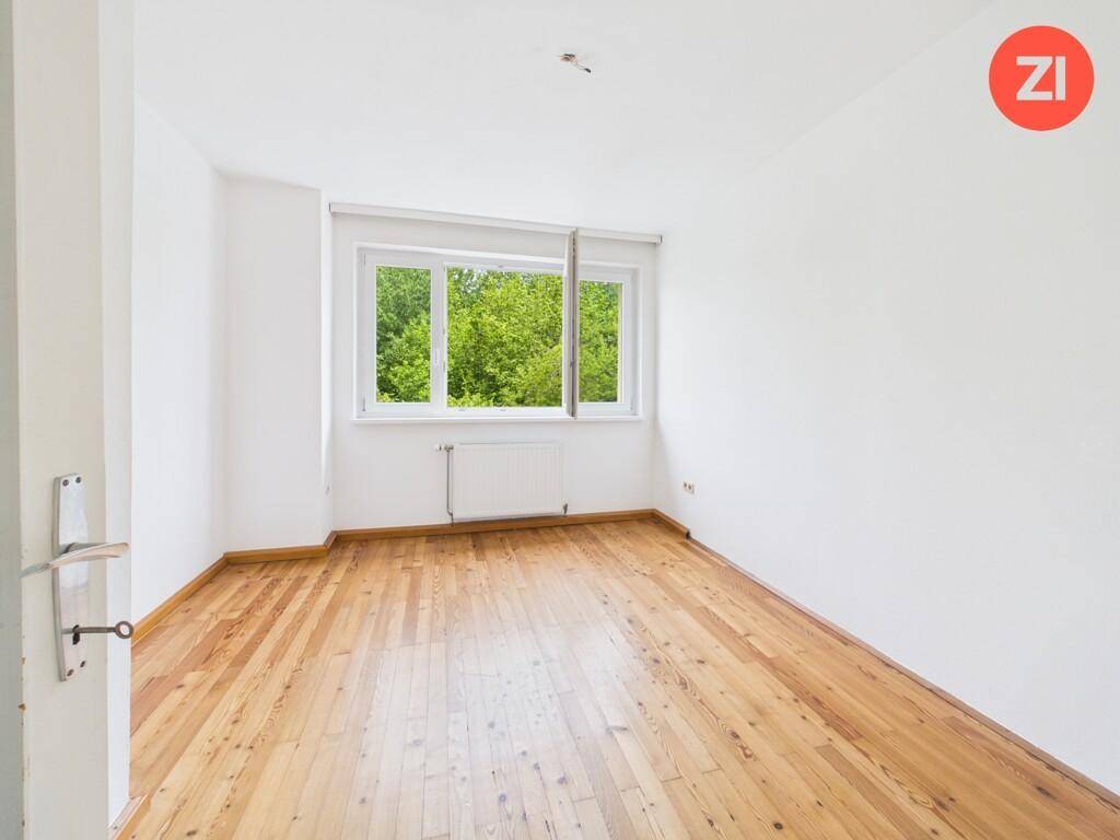 Leeres Zimmer mit Holzboden und großem Fenster, das einen Blick ins Grüne bietet.