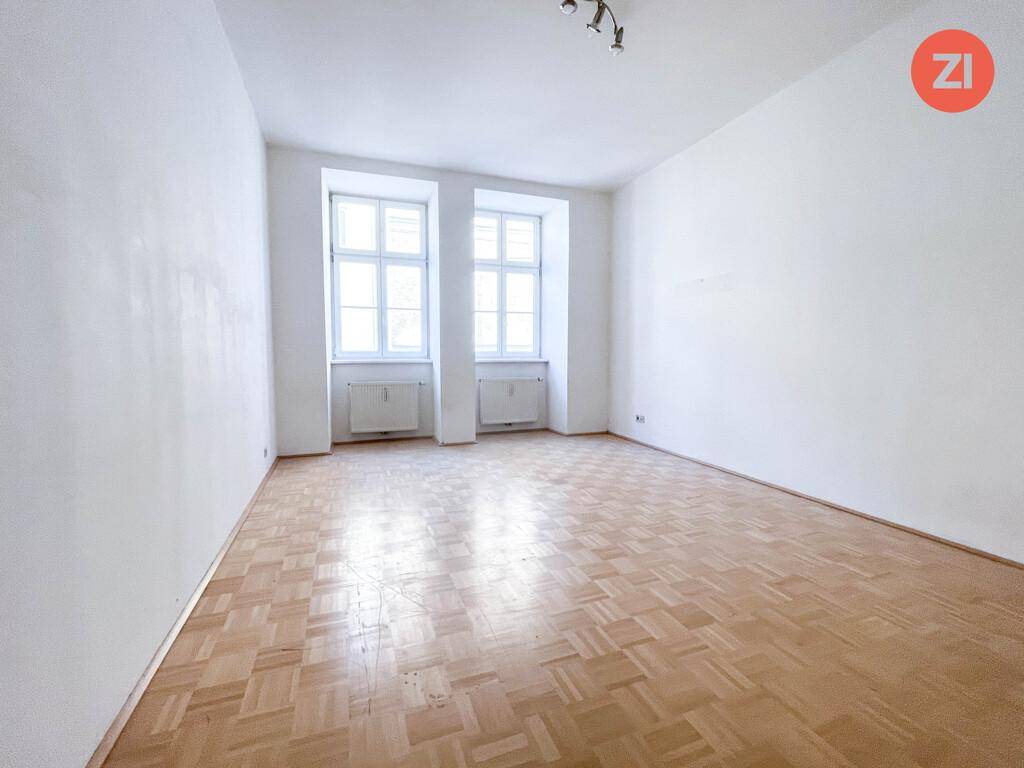 Helles Zimmer mit Fischgrätparkett und zwei großen Fenstern, die viel Tageslicht hereinlassen.