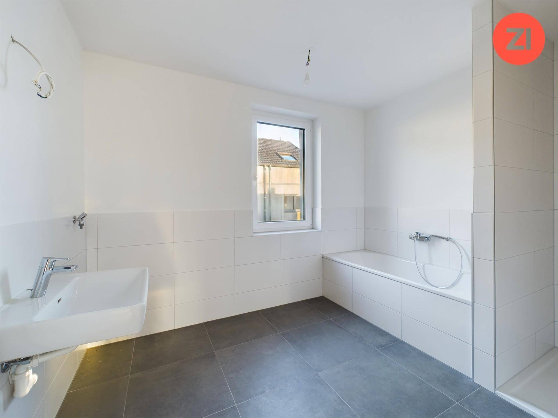 Modernes Badezimmer mit Badewanne, Waschbecken, Fenster und grauen Fliesen auf dem Boden.