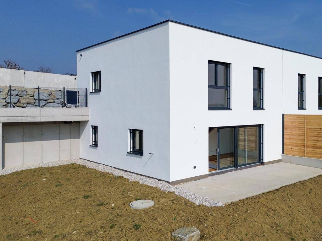 Moderne weiße Hausfassade mit großen Fenstern und Schiebetüren, umgeben von einem unfertigen Gartenbereich.