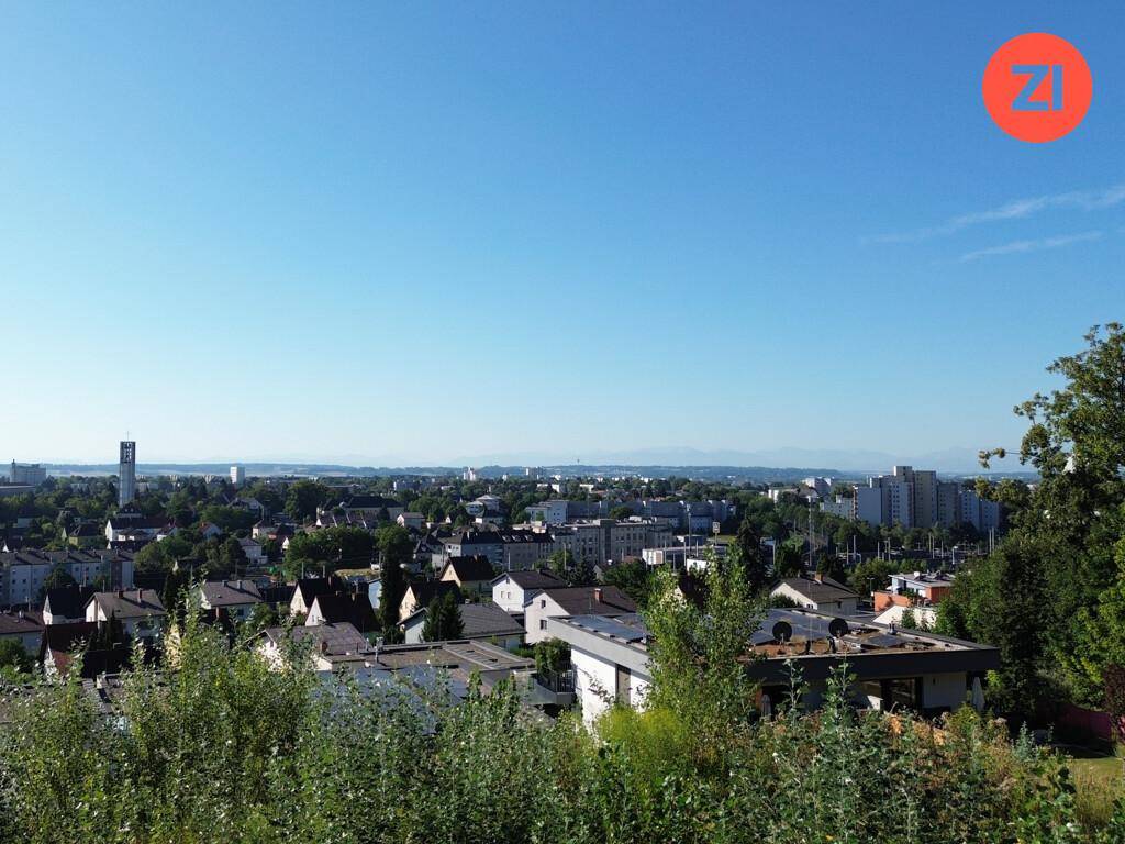 Weitläufiger Blick über eine Stadt mit Wohngebäuden und bewaldeten Hügeln im Vordergrund.