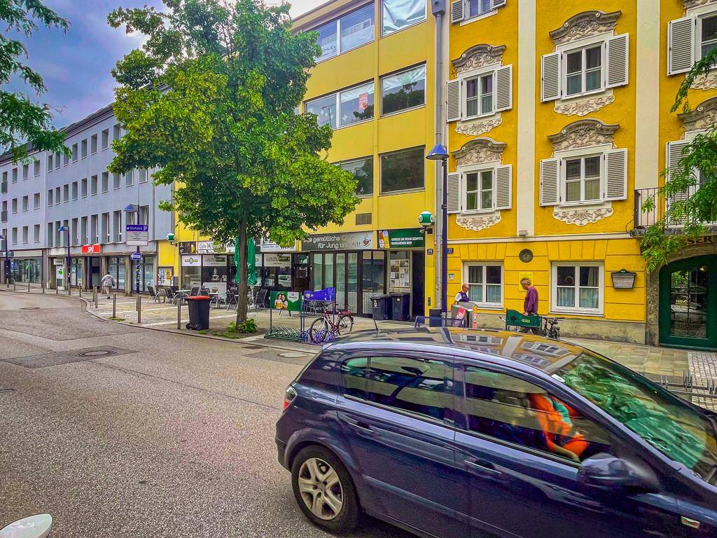 Städtische Straßenszene mit Gebäuden in verschiedenen Farben, einem geparkten Auto und Bäumen.